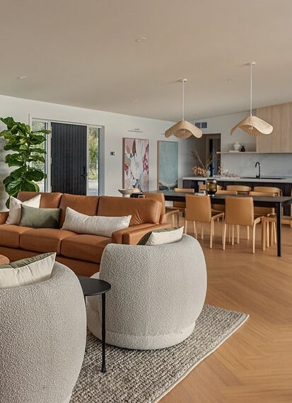 A modern open-plan living room with a tan leather sofa, two round white chairs, a rug, dining table, kitchen area, and large indoor plant near the entrance.