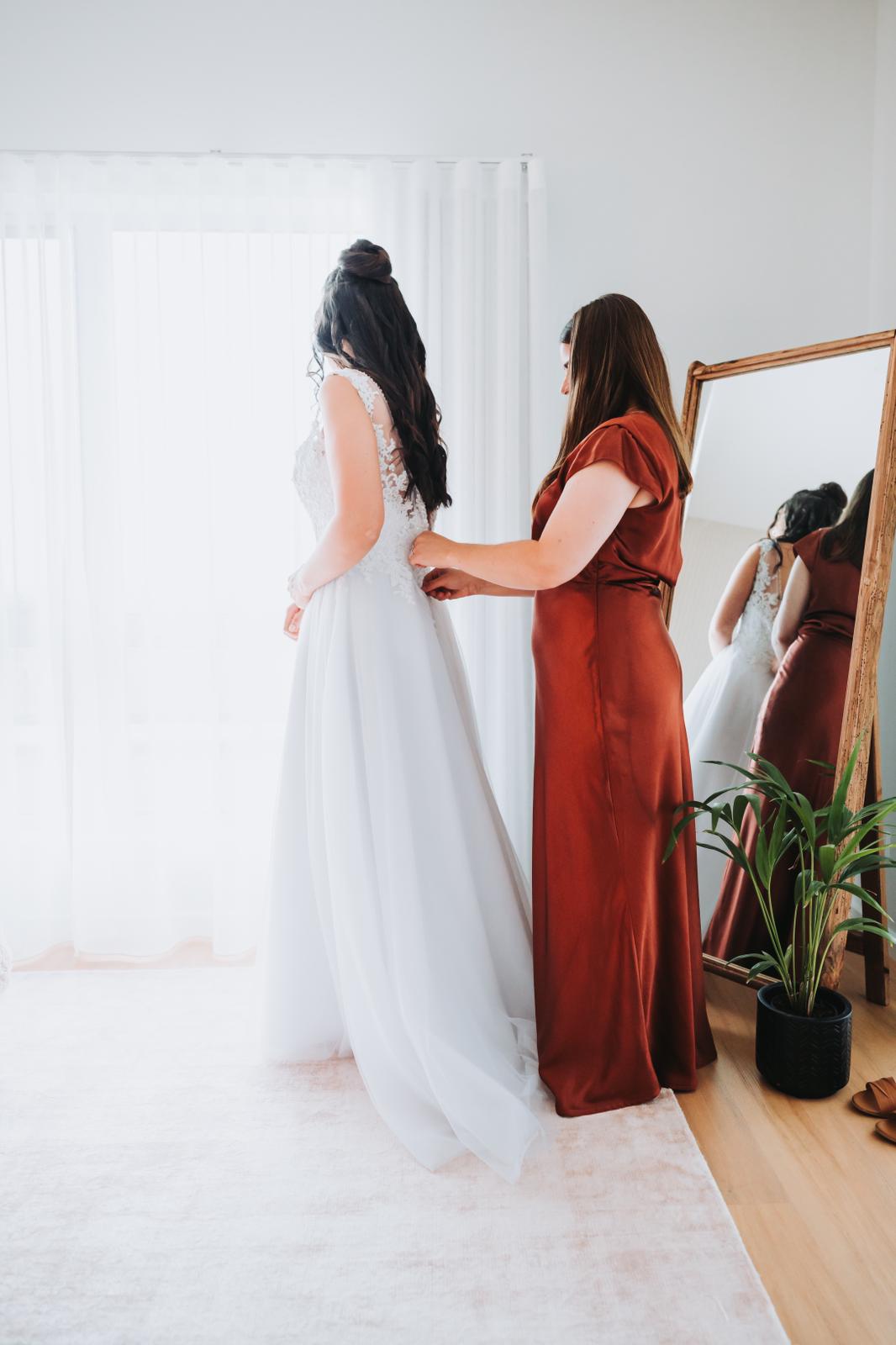 A woman in a rust-colored dress helps another woman in a white wedding gown adjust her dress near a window, with a large mirror and potted plant nearby.